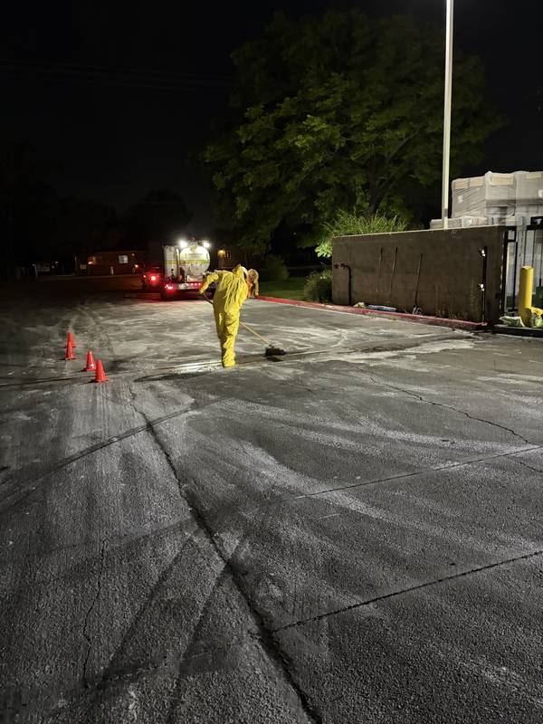Crew member cleaning a paved lot at night with a recovery truck in the background